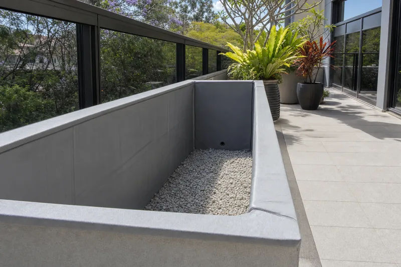 A rendered concrete planter box on an apartment terrace, emptied of soil, with waterproofing membrane dressed up the inside faces and over the lip and drainage gravel in the base — illustrative example