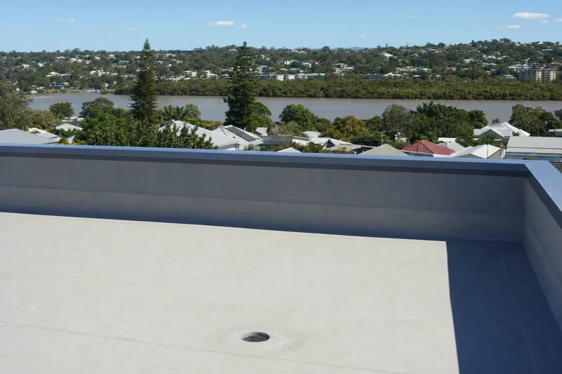 A rooftop deck finished in pale grey trafficable membrane turned up a low parapet under metal capping, with a roof drain outlet in the deck and the river and its mangrove fringe beyond — illustrative example