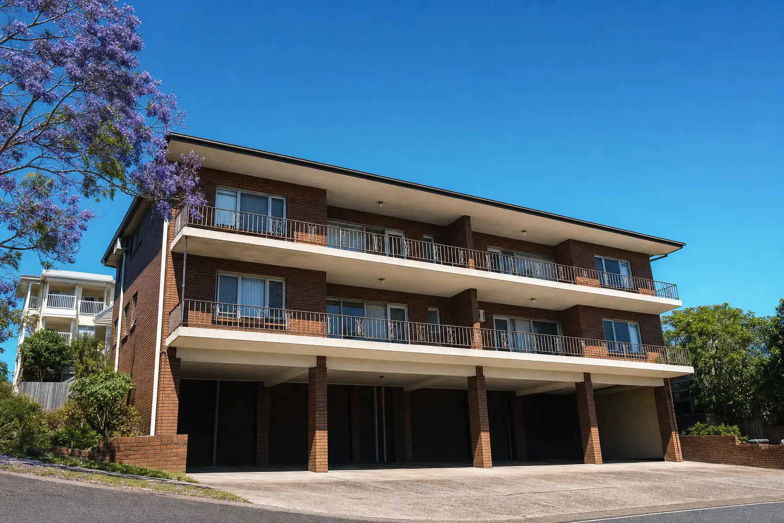 Stacked concrete balconies on a three-storey red-brick walk-up apartment block on a sloping inner-Brisbane street, jacaranda in bloom above — illustrative example