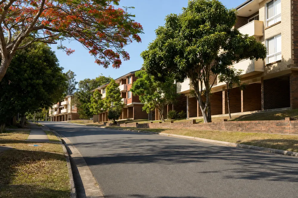 A quiet street of 1960s–80s low-rise brick walk-up apartment blocks with concrete balconies and empty carport bays, poinciana and mango trees in the front yards — illustrative example
