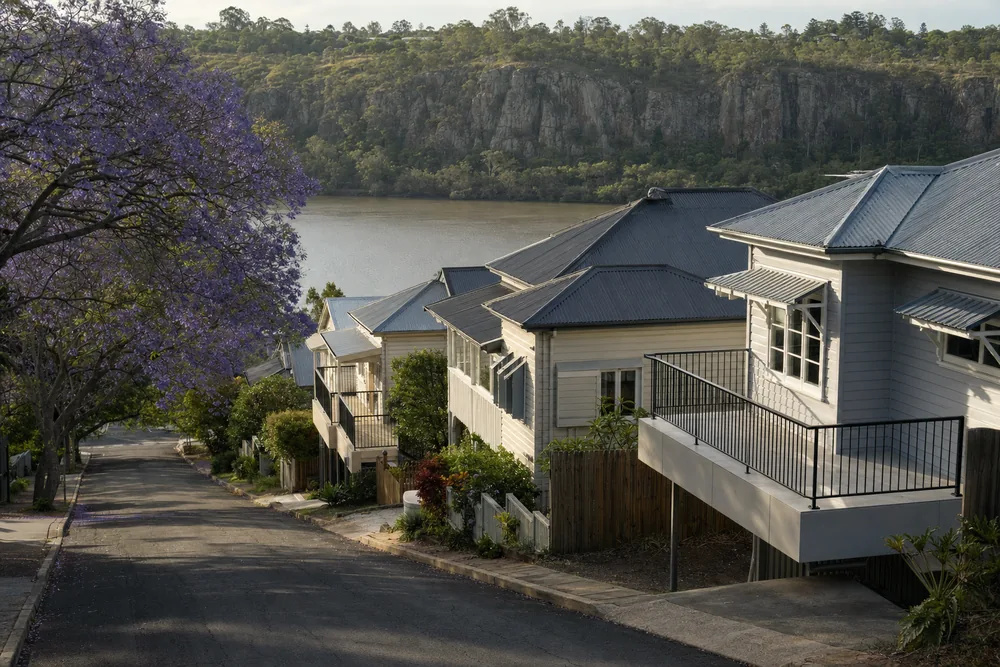 High-set timber Queenslanders with tin roofs stepping down a steep street, one carrying a later concrete deck, the river and a rock cliff beyond — illustrative example